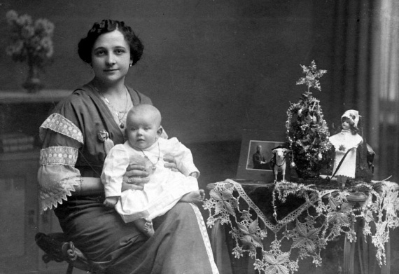 Aprócska fenyőfa mellett fényképezkedett az anyuka, ölében a babával: a fa alá játék kutya és játék baba is került.
                        (1906)