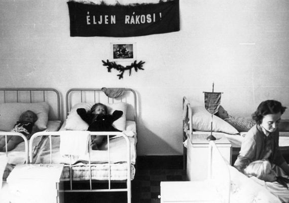 
                        	Balatonlellén ilyen rácsos ágyakban aludtak a gyerkőcök 1952-ben. A háttérben a politikai üzenet árulkodik a tábor jellegéről.